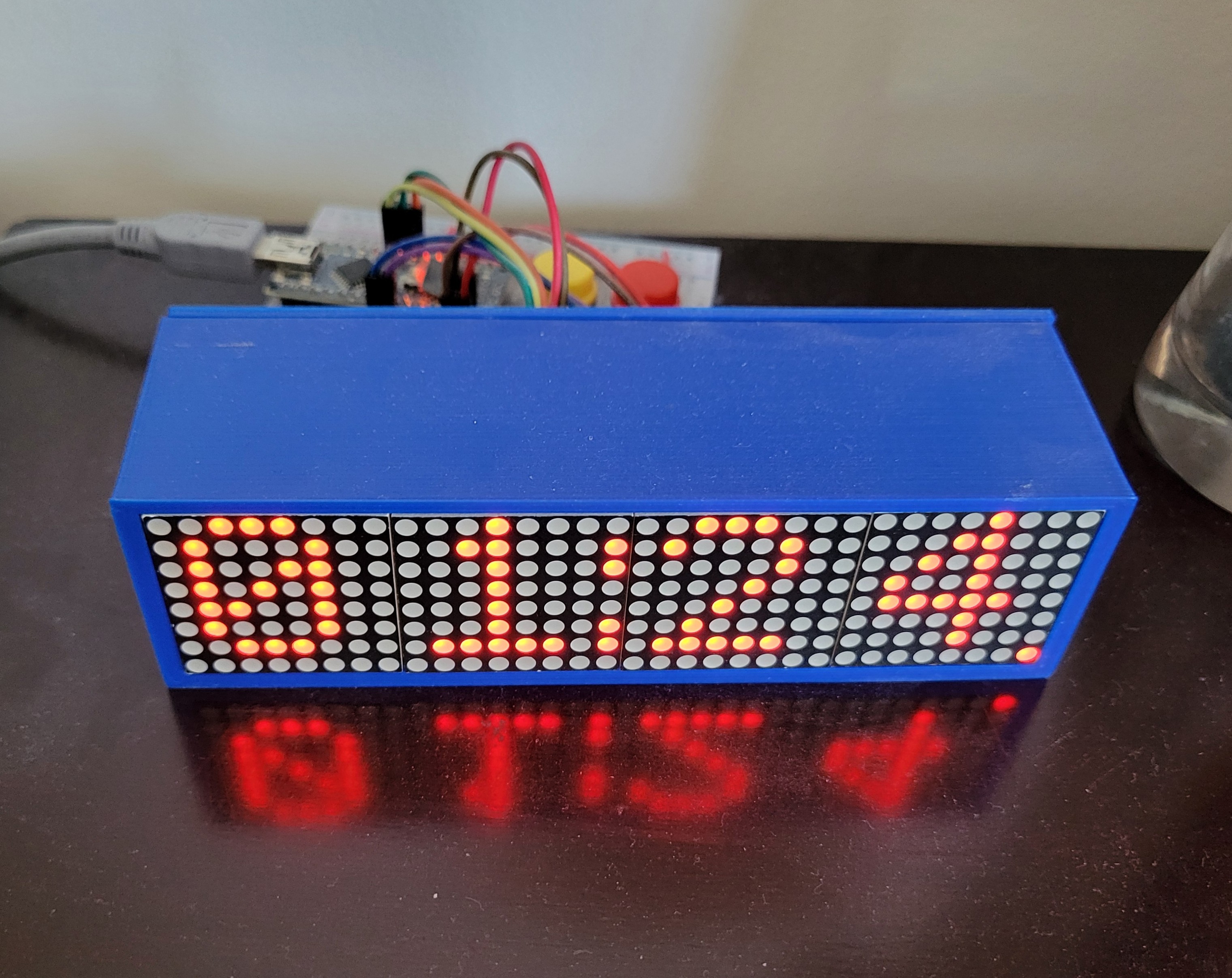 A simple clock with a LED matrix with the current time in a blue case. An Arduino Nano and other components are in the background.